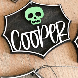 K&ouml;nnte beinhalten: Ein schwarzes Holzschild f&uuml;r Halloween mit einem wei&szlig;en Spinnennetz-Design und dem Namen "Cooper" in wei&szlig;. Ein gr&uuml;ner Sch&auml;del befindet sich oben in der Mitte des Schildes.