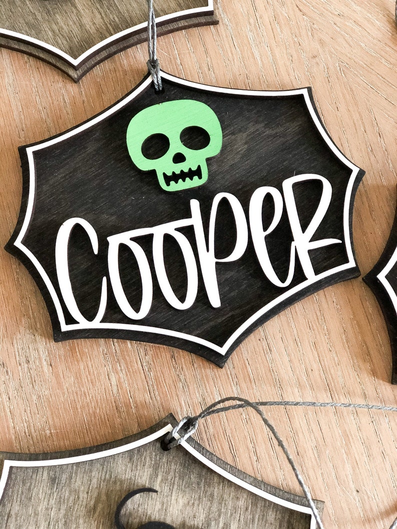 K&ouml;nnte beinhalten: Ein schwarzes Holzschild f&uuml;r Halloween mit einem wei&szlig;en Spinnennetz-Design und dem Namen "Cooper" in wei&szlig;. Ein gr&uuml;ner Sch&auml;del befindet sich oben in der Mitte des Schildes.