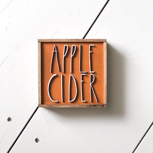 May include: A small wooden sign with a light orange background and a brown frame. The sign says "APPLE CIDER" in white lettering.
