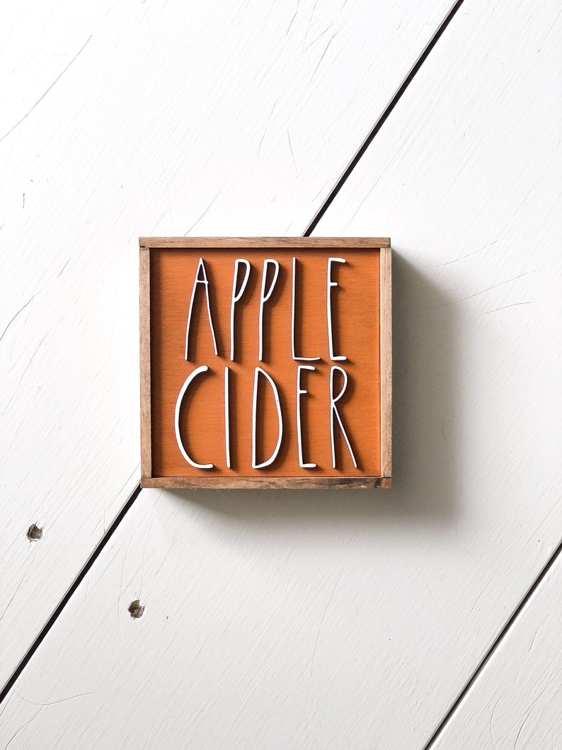May include: A small wooden sign with a light orange background and a brown frame. The sign says "APPLE CIDER" in white lettering.