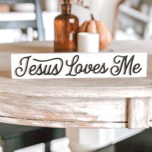 Peut inclure: Une pancarte en bois blanc avec une inscription en cursive noire qui dit "Jesus Loves Me". La pancarte est posée sur une table en bois clair.