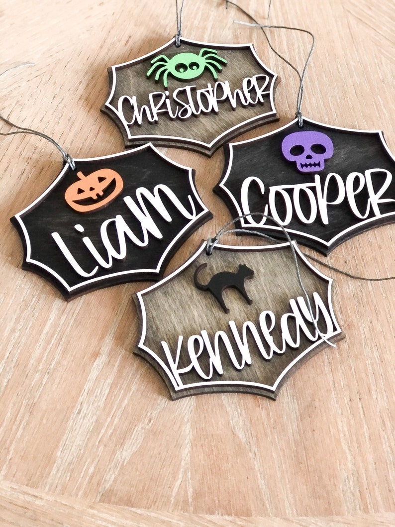 K&ouml;nnte beinhalten: Vier h&ouml;lzerne Halloween-Anh&auml;nger in Form von Spinnweben mit wei&szlig;em Rand. Jeder Anh&auml;nger ist mit einem anderen Design verziert: einer Spinne, einem Sch&auml;del, einem K&uuml;rbis und einer schwarzen Katze. Die Anh&auml;nger sind mit den Namen Christopher, Cooper, Liam und Kennedy personalisiert.