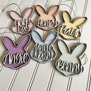 May include: Six wooden bunny-shaped ornaments with different names written on them. The names are Abby Kay, Henry, Paul, Uncle Nasty, Angie, and Benny. The ornaments are painted in various pastel colors.