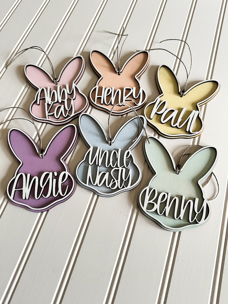 May include: Six wooden bunny-shaped ornaments with different names written on them. The names are Abby Kay, Henry, Paul, Uncle Nasty, Angie, and Benny. The ornaments are painted in various pastel colors.