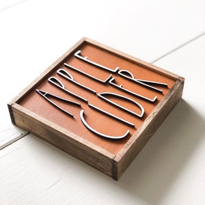 May include: A wooden sign with a brown background and a white wood frame. The sign says "Apple Cider" in white letters.