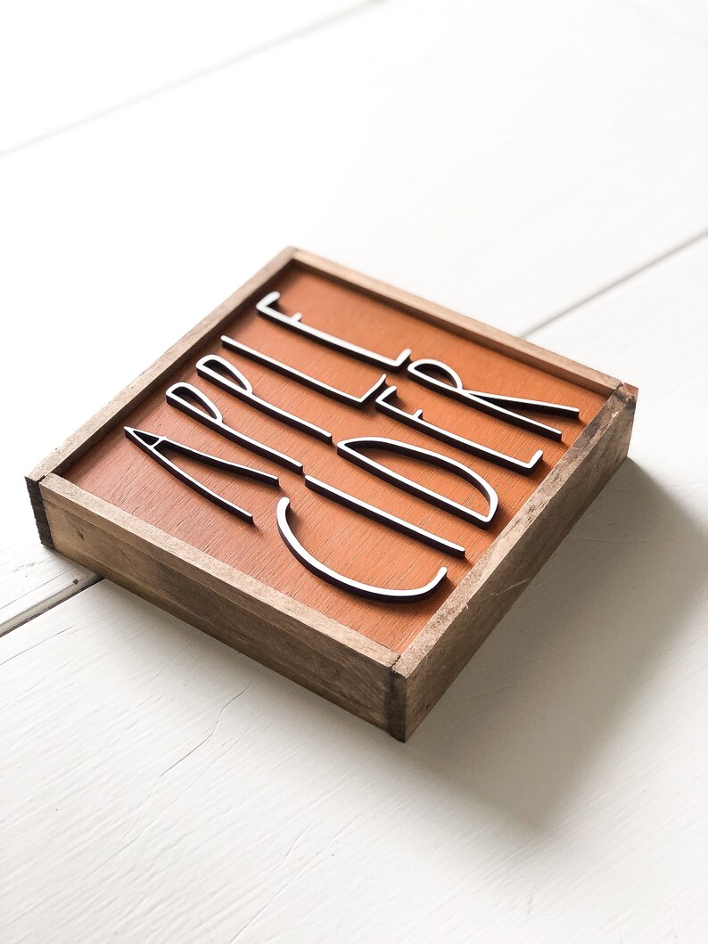 May include: A wooden sign with a brown background and a white wood frame. The sign says "Apple Cider" in white letters.