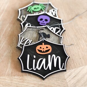 K&ouml;nnte beinhalten: Ein Set aus f&uuml;nf schwarzen Halloween-Anh&auml;ngern mit verschiedenen Designs. Die Anh&auml;nger sind in Form einer Spinne, eines Sch&auml;dels, einer Katze, eines K&uuml;rbisses und des Namens "Liam" gestaltet.