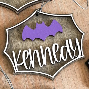 K&ouml;nnte beinhalten: H&ouml;lzerner Halloween-Anh&auml;nger mit einer Fledermaus-Silhouette und dem Namen "Kennedy" in wei&szlig;en Buchstaben. Der Anh&auml;nger ist braun mit einem wei&szlig;en Rand und einer lila Fledermaus.