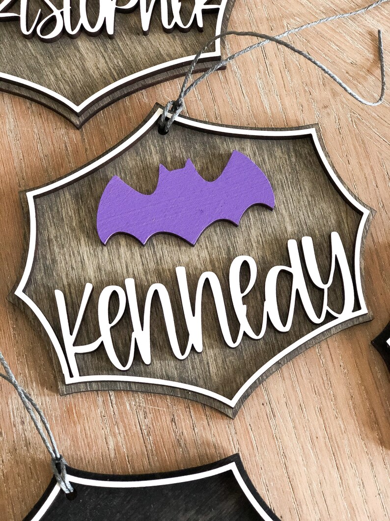 K&ouml;nnte beinhalten: H&ouml;lzerner Halloween-Anh&auml;nger mit einer Fledermaus-Silhouette und dem Namen "Kennedy" in wei&szlig;en Buchstaben. Der Anh&auml;nger ist braun mit einem wei&szlig;en Rand und einer lila Fledermaus.