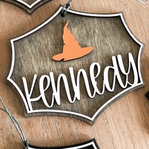 K&ouml;nnte beinhalten: Ein h&ouml;lzerner Halloween-Anh&auml;nger mit einem Hexenhut und dem Namen "Kennedy" in wei&szlig;. Der Anh&auml;nger ist braun mit einem wei&szlig;en Rand und einer Schnur zum Aufh&auml;ngen.