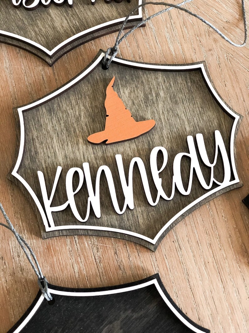 K&ouml;nnte beinhalten: Ein h&ouml;lzerner Halloween-Anh&auml;nger mit einem Hexenhut und dem Namen "Kennedy" in wei&szlig;. Der Anh&auml;nger ist braun mit einem wei&szlig;en Rand und einer Schnur zum Aufh&auml;ngen.