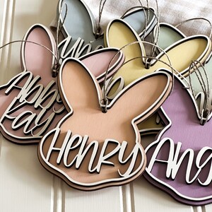 May include: Five pastel colored wooden bunny shaped ornaments with the names Henry, Abby, Willow, Angie, and a fourth name that is unclear. The ornaments are hanging from twine.