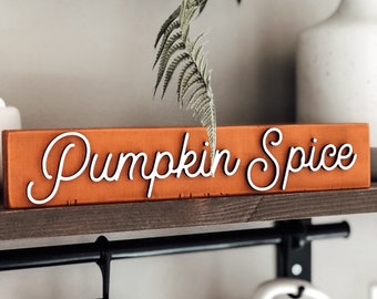 May include: A wooden sign with the text "Pumpkin Spice" in white cursive lettering on an orange background. The sign is on a shelf with a black metal rod and hooks hanging from it. There are two mugs hanging from the hooks, one black and one white.