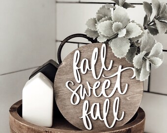 May include: A wooden tray holds a round sign with the words "fall sweet fall" in white script. A white house-shaped object and a black cube sit on the tray. Faux greenery adds a decorative touch to this fall-themed decor.