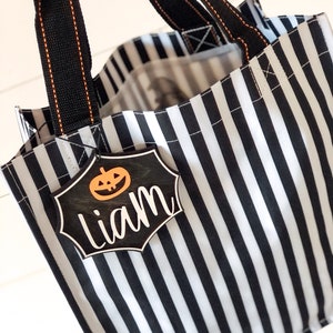 K&ouml;nnte beinhalten: Schwarze und wei&szlig;e gestreifte Halloween-Trick-or-Treat-Tasche mit schwarzen Griffen und einem schwarzen und wei&szlig;en K&uuml;rbis-f&ouml;rmigen Anh&auml;nger mit der Aufschrift "Liam".