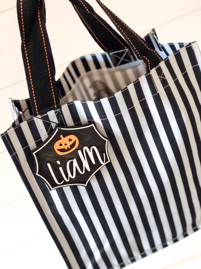 K&ouml;nnte beinhalten: Schwarze und wei&szlig;e gestreifte Halloween-Trick-or-Treat-Tasche mit schwarzen Griffen und einem schwarzen und wei&szlig;en K&uuml;rbis-f&ouml;rmigen Anh&auml;nger mit der Aufschrift "Liam".