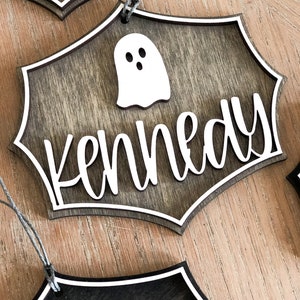 K&ouml;nnte beinhalten: Ein h&ouml;lzernes Halloween-Schild mit einem Geister-Ausschnitt und dem Namen "Kennedy" in wei&szlig;en Buchstaben.