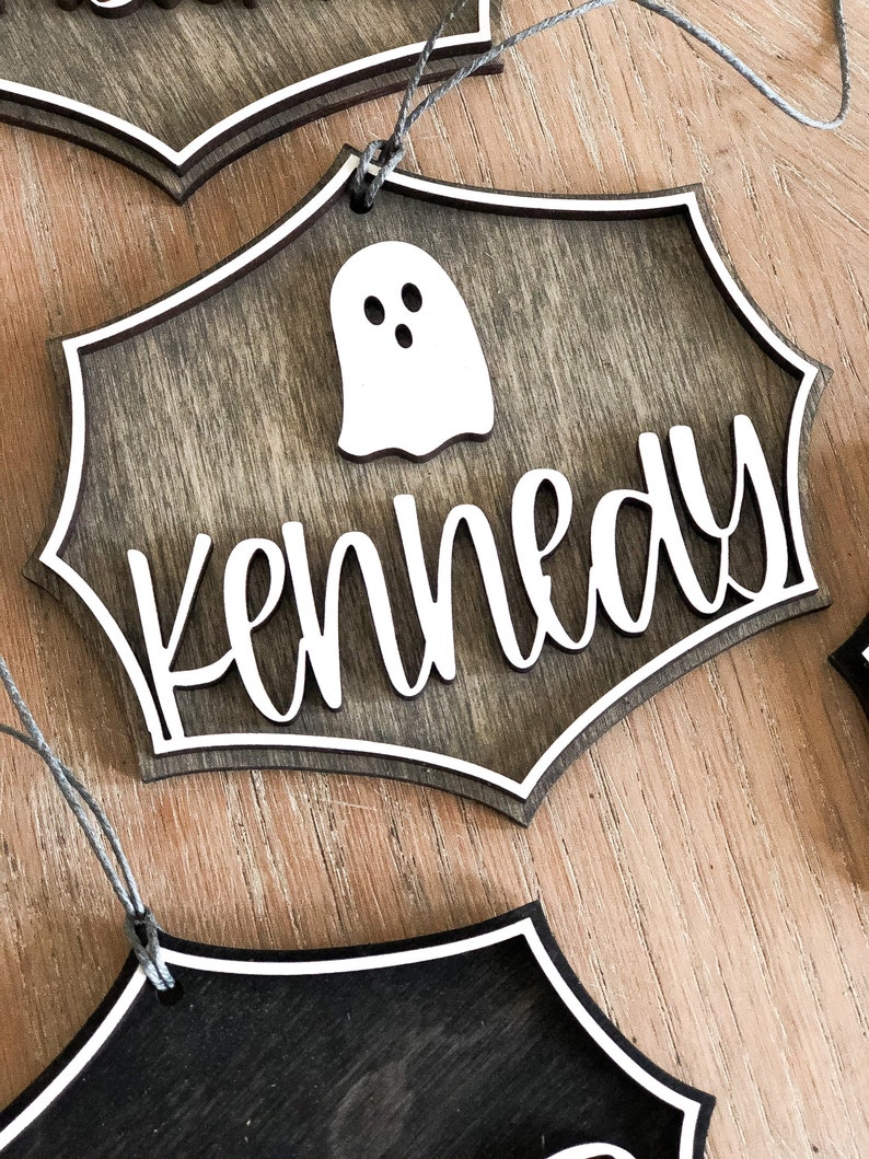 K&ouml;nnte beinhalten: Ein h&ouml;lzernes Halloween-Schild mit einem Geister-Ausschnitt und dem Namen "Kennedy" in wei&szlig;en Buchstaben.