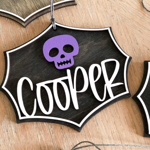 K&ouml;nnte beinhalten: Ein schwarzer Holz-Halloween-Anh&auml;nger mit einem wei&szlig;en Spinnennetz-Design und dem Namen "Cooper" in wei&szlig;. Ein lila Sch&auml;del ist oben am Anh&auml;nger befestigt.