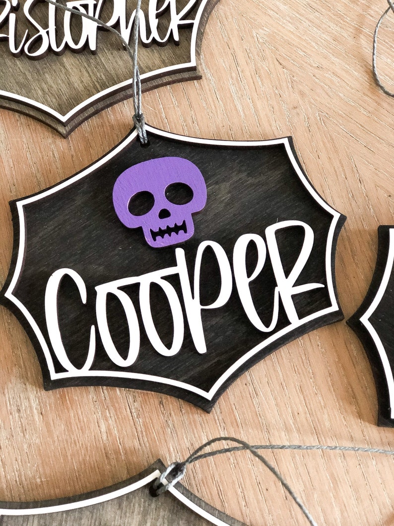 K&ouml;nnte beinhalten: Ein schwarzer Holz-Halloween-Anh&auml;nger mit einem wei&szlig;en Spinnennetz-Design und dem Namen "Cooper" in wei&szlig;. Ein lila Sch&auml;del ist oben am Anh&auml;nger befestigt.