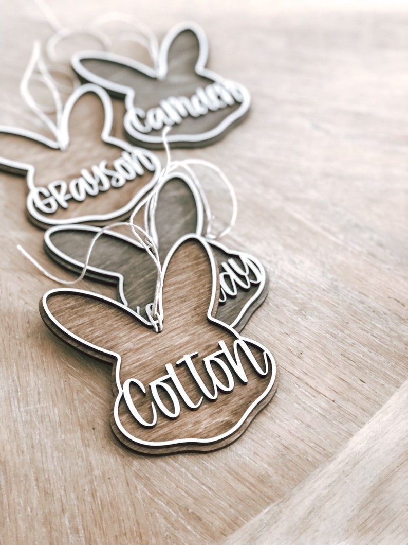 May include: Four wooden bunny-shaped ornaments with the names Grayson, Camden, Lily, and Colton written on them. The ornaments are hanging from twine.
