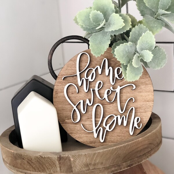 May include: A round wooden sign with the words "home sweet home" written in white cursive lettering. The sign is sitting on a tiered wooden tray with a small black and white house figurine.