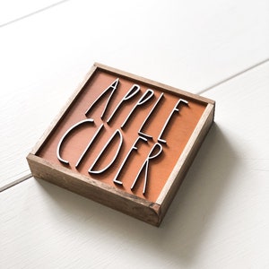 May include: A wooden sign with a brown background and the words "APPLE CIDER" in black lettering.