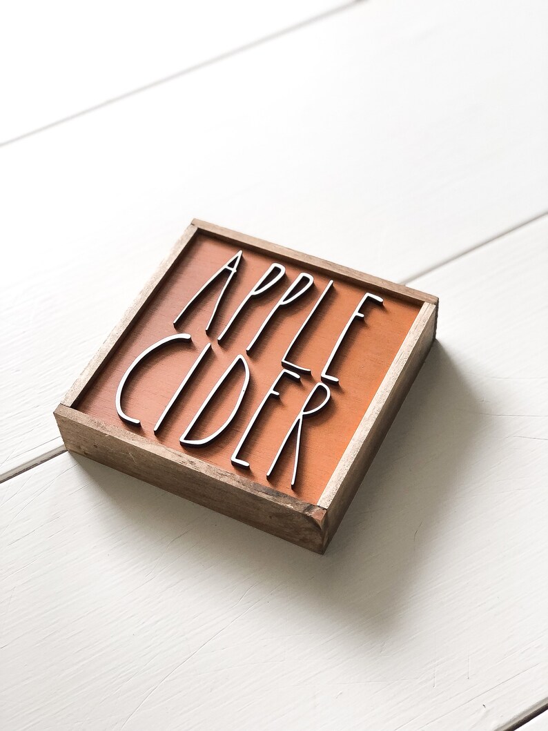 May include: A wooden sign with a brown background and the words "APPLE CIDER" in black lettering.