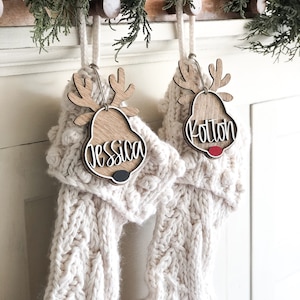 Peut inclure: Deux bas en tricot blanc avec des ornements de renne en bois. Les ornements portent les noms "Jessica" et "Kolton".