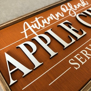Peut inclure: Une enseigne en bois avec un fond brun et du texte blanc. L'enseigne indique "Autumn Blend Apple Cider" et "Serving".