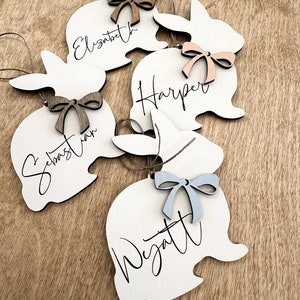 Peut inclure: Cinq décorations de lapin en bois blanc avec des noms écrits en calligraphie noire. Les lapins ont des nœuds de différentes couleurs attachés à eux : marron, pêche, bleu et deux nœuds marrons.