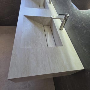 Puede incluir: Un tocador doble de piedra beige claro con una textura veteada natural. Cuenta con dos lavabos rectangulares integrados con lados inclinados y una cubierta de drenaje central. Dos grifos de n&iacute;quel cepillado est&aacute;n montados en la encimera.