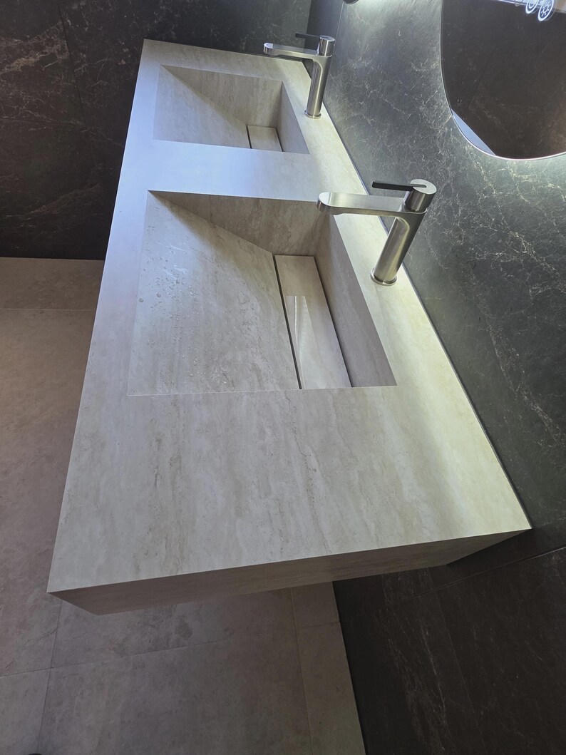 Puede incluir: Un tocador doble de piedra beige claro con una textura veteada natural. Cuenta con dos lavabos rectangulares integrados con lados inclinados y una cubierta de drenaje central. Dos grifos de n&iacute;quel cepillado est&aacute;n montados en la encimera.