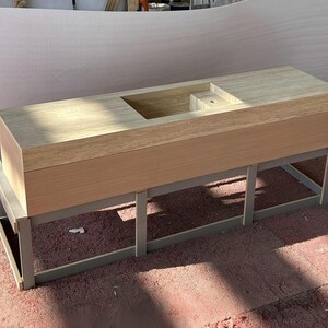 Puede incluir: Un mueble de lavabo rectangular con una encimera de piedra travertino de color beige claro, que incluye un lavabo rectangular integrado y un orificio para el grifo. La base es un mueble de madera clara sobre una estructura de metal gris.