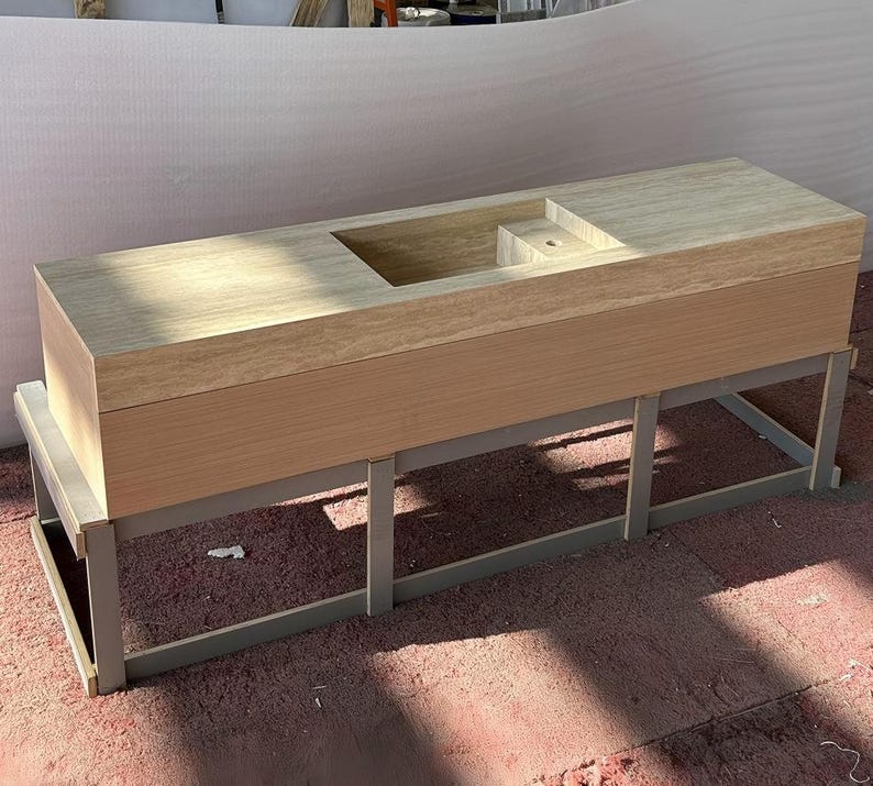 Puede incluir: Un mueble de lavabo rectangular con una encimera de piedra travertino de color beige claro, que incluye un lavabo rectangular integrado y un orificio para el grifo. La base es un mueble de madera clara sobre una estructura de metal gris.