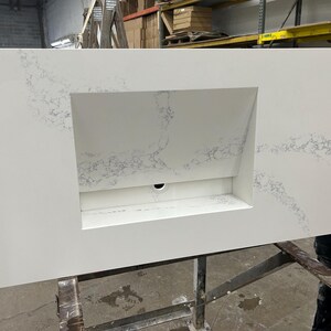 May include: A large, rectangular white countertop with a built-in rectangular sink basin. The countertop has subtle gray veining throughout. A black drain hole is visible in the sink basin. The countertop is being held by a person.