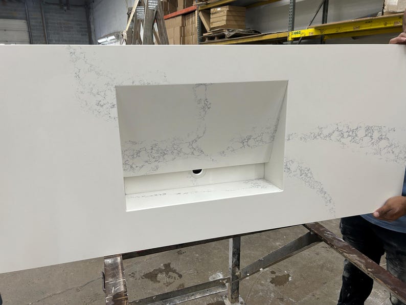 May include: A large, rectangular white countertop with a built-in rectangular sink basin. The countertop has subtle gray veining throughout. A black drain hole is visible in the sink basin. The countertop is being held by a person.