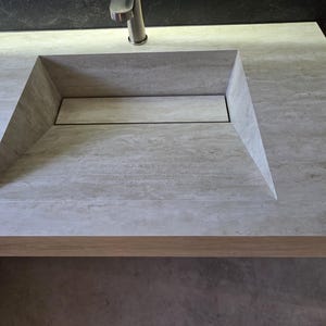 Puede incluir: Un lavabo de piedra gris claro integrado en una encimera a juego con una cubeta rectangular y una tapa de desag&uuml;e lineal oculta. Un grifo de metal cepillado est&aacute; colocado en la parte central trasera del lavabo de superficie texturizada.