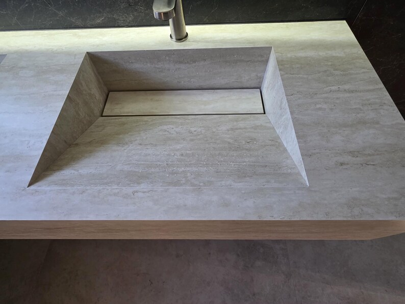 Puede incluir: Un lavabo de piedra gris claro integrado en una encimera a juego con una cubeta rectangular y una tapa de desag&uuml;e lineal oculta. Un grifo de metal cepillado est&aacute; colocado en la parte central trasera del lavabo de superficie texturizada.