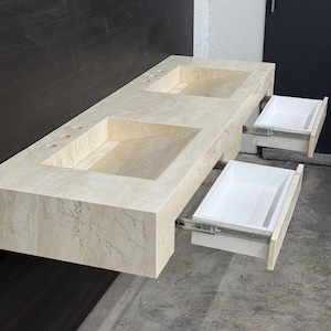 May include: A beige marble double sink vanity with two rectangular basins. Two white drawers are partially extended. The vanity is mounted on a dark brown wall. The overall design is modern and minimalist.