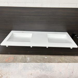 May include: A white rectangular bathroom vanity countertop with two integrated sinks. The countertop has a clean, modern design with a smooth surface and is made of a solid, durable material. The countertop is designed for a double sink setup.