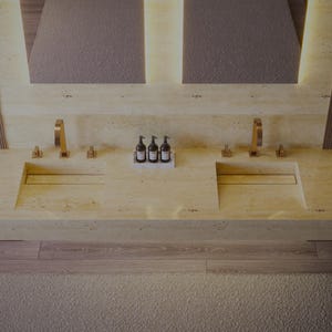 Puede incluir: Un tocador doble de piedra beige claro con dos lavabos rectangulares integrados, dos grifos arqueados dorados con manijas separadas y una peque&ntilde;a bandeja blanca que sostiene tres botellas con dosificador marr&oacute;n oscuro en el centro.