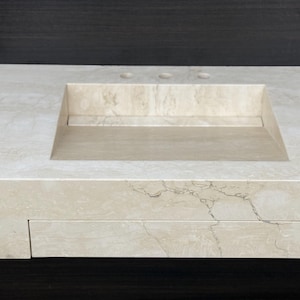 Puede incluir: Un lavabo rectangular de m&aacute;rmol beige con una superficie pulida y lisa que presenta vetas naturales. El seno est&aacute; integrado en la losa de piedra, la cual incluye tres orificios preperforados para la grifer&iacute;a en el borde posterior.