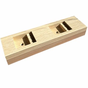 Puede incluir: Un bloque de madera rectangular de color claro que presenta dos compartimentos cuadrados empotrados. Cada compartimento contiene un peque&ntilde;o orificio circular en la base y una ranura vertical en el lado interior, mostrando la veta natural de la madera.