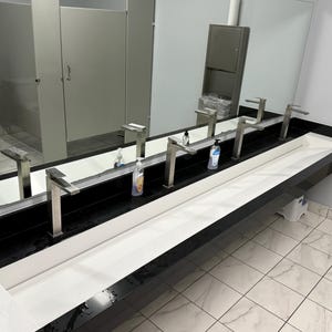 May include: A public restroom with multiple sinks and mirrors. The sinks are stainless steel with modern faucets. Soap dispensers are placed on the countertop. The countertop is black with a white sink basin. The floor is tiled.