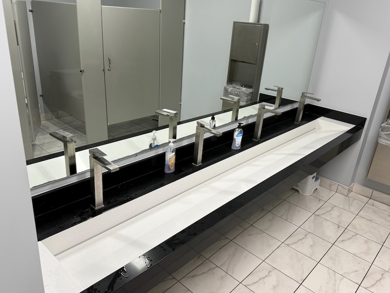 May include: A public restroom with multiple sinks and mirrors. The sinks are stainless steel with modern faucets. Soap dispensers are placed on the countertop. The countertop is black with a white sink basin. The floor is tiled.