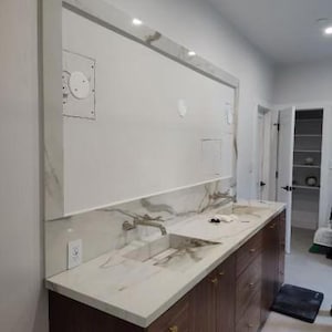 May include: A modern bathroom vanity with a marble countertop and dark wood cabinets. The vanity features two sinks, gold hardware, and a large mirror with integrated lighting. The walls are painted white, and the room has a minimalist design.