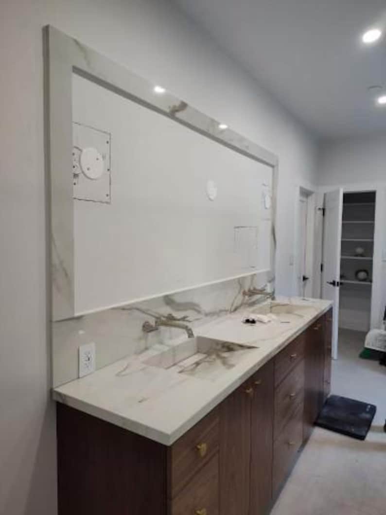 May include: A modern bathroom vanity with a marble countertop and dark wood cabinets. The vanity features two sinks, gold hardware, and a large mirror with integrated lighting. The walls are painted white, and the room has a minimalist design.
