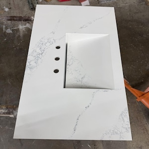 May include: A white countertop with a built-in rectangular sink and three faucet holes. The countertop has subtle gray veining throughout. The sink is a bright white, and the overall design is modern and clean.