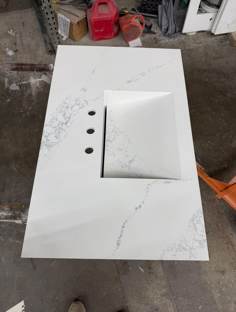 May include: A white countertop with a built-in rectangular sink and three faucet holes. The countertop has subtle gray veining throughout. The sink is a bright white, and the overall design is modern and clean.